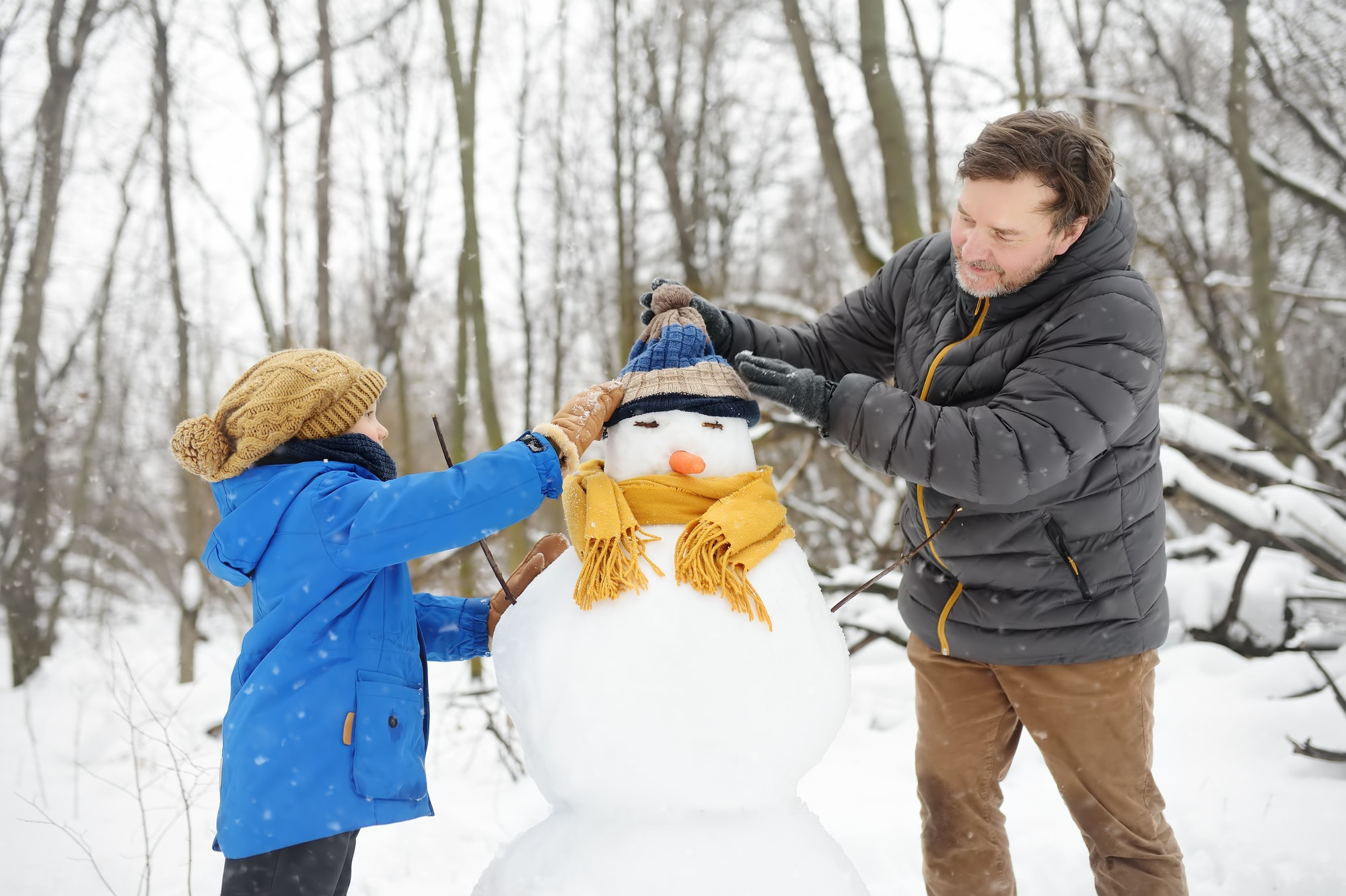 Little boy with his father building snowman in snowy park. Dad and son tied a scarf for snowman.